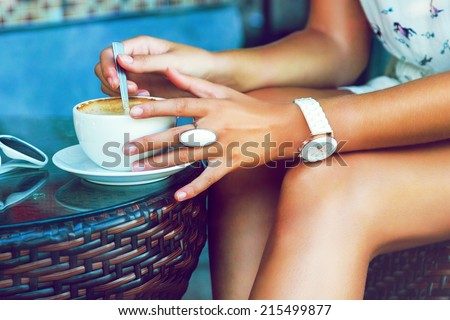Close up image of tan stylish woman shaking her morning coffee, wearing summer trendy dress and accessorizes , siring alone at cafe. Bright colors.