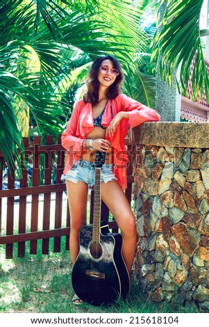 Young sexy beautiful woman posing at palms garden in hot summer sunny day with acoustic guitar, wearing trendy boho styled clothes and sunglasses. Bright colors .