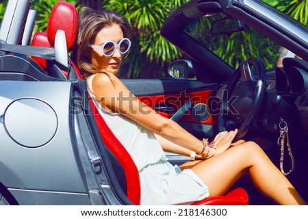 Stylish pretty tan woman riding luxury cabriolet car, enjoy her adventures in sunny day at hot topical country, wearing relaxed beach dress and sunglasses.