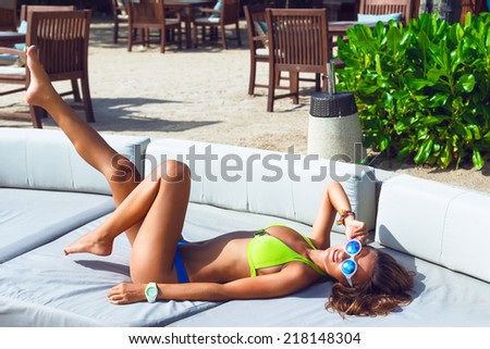 Outdoor portrait of pretty slim tanned woman, having fun alone, relaxed on big white sofa at luxury resort on hot tropical counter, wearing stylish neon bikini and sunglasses, put her leg in the air.