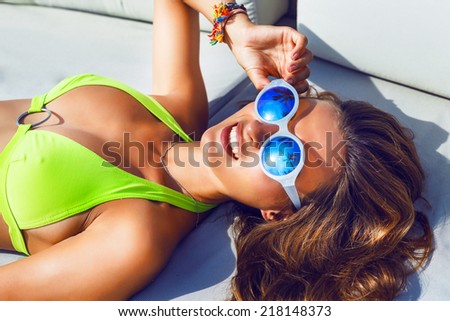 Close up outdoor fashion portrait of young woman with perfect bronze tanned skin, laying and herring sunbathe, wearing bright bikini and blue mirrored sunglasses with reflection of palms on it.