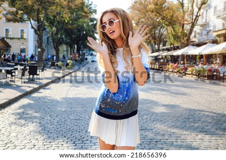 Funny playful portrait of pretty blonde girl, having fun at the street at european city  have surprised emotions ant put her hands in the air. wearing bright retro clothiers. Nice sunny fall day.