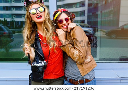 Lifestyle bright urban portrait of two bear fiends. Two pretty girls wearing swag hipster neon hats and sunglasses, hugs and having great time together, city window background.