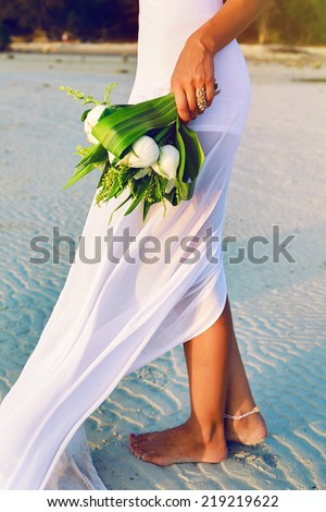 Outdoor fashion image of bride posing alone at exotic beach at sunset with beautiful wedding lotus bouquet, have nice simple trendy white dress.