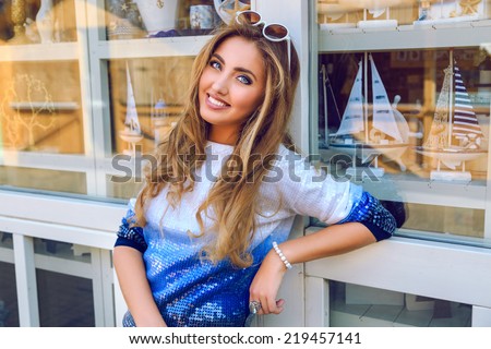 Young pretty smiling girl posing near souvenir shop with boats and sailors goods, wearing ombre sweater, have natural make up and fluffy curled hairstyle. Enjoy nice day and shopping.