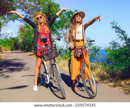 Outdoor sunny fashion portrait of two pretty funny girls, having fun together and going crazy, riding vintage hipster bikes, waring vintage clothes hats and sunglasses. Positive mood.