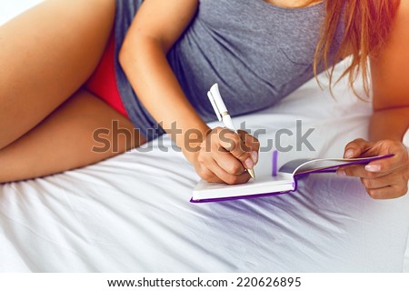 Close up lifestyle image of young woman laying on the bed and making important notes to her diary. Bright colors.