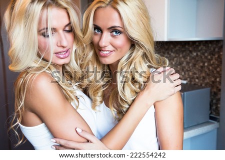 Close up lifestyle indoor fresh fashion portrait of two pretty smiling blonds. Two best friends hugs at kitchen, have fun and happy free time together.