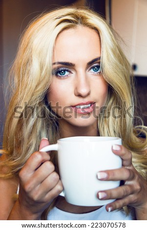 Indoor lifestyle portrait of pretty blonde fresh woman posing at home in the morning, and drinking tea from big white cup.