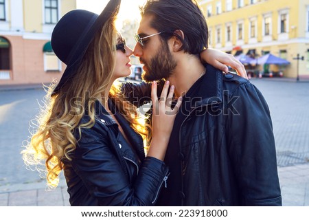 Fashion outdoor sensual romantic portrait of beautiful young couple hugs and kissing on the stereo of european city. Wearing trendy fall black outfits, rock n roll style. Bright evening sunlight.