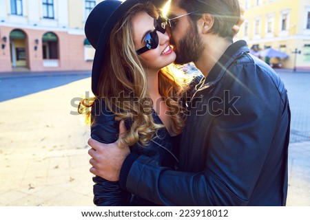 Young handsome stylish guy listing her pretty blonde girlfriend, romantic sensual couple hugs on the street of european city at bright evening sunlight, rock n roll styled clothes and sunglasses.