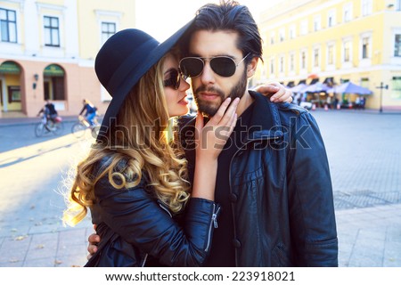 Young beautiful sensual blonde girl kissing her handsome stylish boyfriend. Pretty romantic couple wearing leather jackets, hat and sunglasses, rock biker style. Bright evening street sunlight.