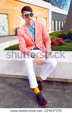 Young stylish hipster man posing at shopping street, wearing bright classic vintage casual look, denim shirt, peach jacket and sunglasses. Outdoor fashion portrait, urban background.