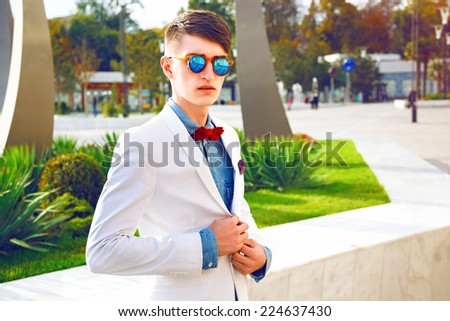 Young stylish hipster man posing at modern city park at nice sunny day, wearing mirrored sunglasses, white jacket, denim shirt and red bow tie. Bright colors.