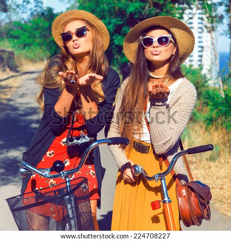 Two pretty young happy girls send you air kiss, wearing vintage stylish clothes hats and sunglasses, having fun together and walking with retro bikes. Fashion outdoor portrait of two sisters.