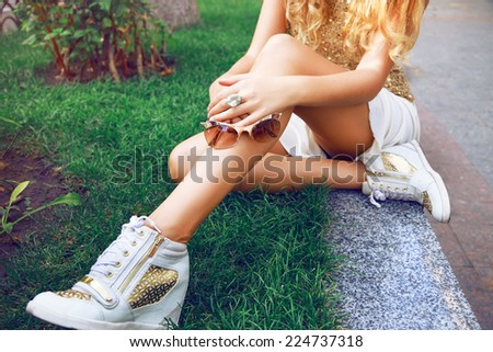 Outdoor fashion image of young slim fit woman sitting on the grass in mini skirt, wearing golden blouse and sneakers, holding sunglasses on her hands.