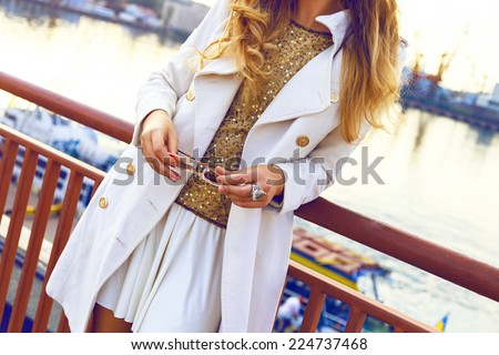 Close up fashion image of woman wearing stylish fall outfit, white mini skirt cashmere coat, golden top and sunglasses, amazing view on sea port, soft evening light.