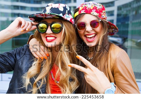 Closeup fashion lifestyle portrait of two pretty best friends girls, wearing bright swag style floral hats, mirrored sunglasses, having fun and make crazy funny faces. Two sisters posing on party.