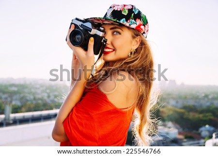 Young sexy smiling photographer woman, holding and making picture on retro vintage camera, wearing swag floral bright cap, amazing view on the city from roof, joy and happiness.