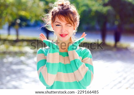 Young happy smiling teen girl winks to you, have positive mod and emotions, wearing bright comfy casual sweater, posing in the park in nice sunny day.