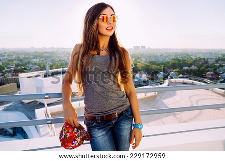 Young pretty fit sportive stylish girl with long brunette hairs posing at the roof, wearing casual outfit swag hat and mirrored sunglasses, amazing view on the city, evening sunlight.