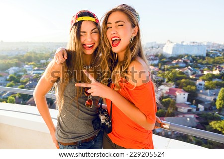 Two crazy smiling happy hipster girls best friends having fun on the roof, making funny playful faces, wearing bright casual outfits hats and sunglasses, holding vintage camera, positive emotions.