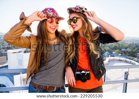 Two sisters posing on the roof, wearing stylish leather jackets swag hats and sunglasses, holding retro camera, ready for shooting, positive emotions. fashion portrait of best friends girls.