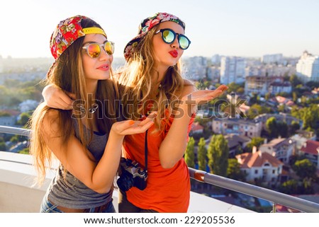 Two pretty fashion girls sending kiss and having fun, wearing bright swag caps and sunglasses, posing at the roof, positive nice emotions and mood, amazing view on city.