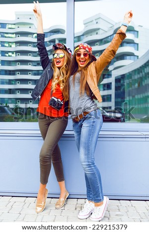 Outdoor urban lifestyle portrait of two pretty girls best friends screaming laughing and put their hands to the air. wearing casual bright clothes hats and sunglasses, happy mood emotions.
