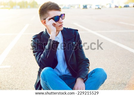 Young handsome man posing at sunny day at city parking, wearing stylish biker leather jacket and sunglasses. Urban style.