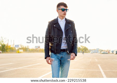 Urban outdoor portrait of young stylish man wearing leather biker jacket and sunglasses, evening sunlight. Posing at countryside parking.