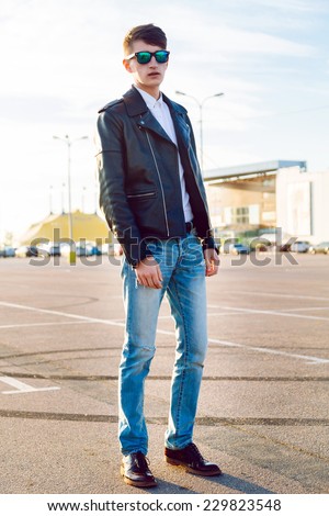 Outdoor fall fashion portrait of stylish man posing at city parking, wearing denim jeans biker black leather jacket and sunglasses.