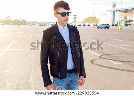 Handsome stylish man posing at countryside parking in evening sunlight, wearing stylish leather rocker jacket and hipster haircut.