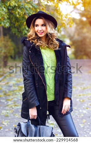 Fashion portrait of pretty young blonde smiling woman wearing trendy coat, vintage hat and neon sweater, posing at countryside park in nice sunny fall autumn day.