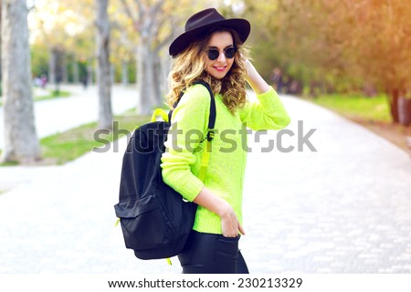 Outdoor fashion image of stylish hipster girl wearing neon sweater sunglasses and vintage hat, walking with back pack on the street in nice sunny fall autumn day.