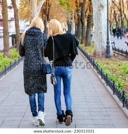 Two pretty friend walking together at city avenue. Fashion fall autumn portrait of stylish blondes wearing trendy cozy casual outfits. Speaking and having fun together.
