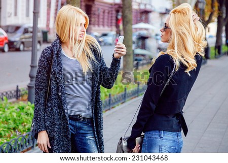 Outdoor lifestyle portrait of two stylish blondes best friends having faun and making photos of each other on smartphone in nice fall autumn day.