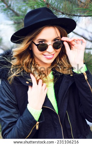 Close up fashion portrait of pretty cheerful girl posing in nice winter day near fir tree, wearing vintage hipster hat and sunglasses, neon sweater and trendy parka. Bright colors happy holidays mood.