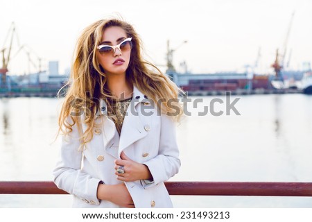 Fall autumn fashion portrait of elegant lady model posing at sea port in evening soft sunlight, wearing extravagant gold sunglasses and white cashmere stylish coat.