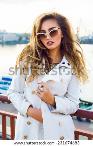 Outdoor evening fall portrait of beautiful sexy blonde woman posing in stylish cashmere coat behind city sea port.