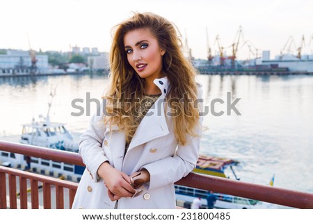 Outdoor fall fashion portrait of sexy elegant lady posing neat sea port in white coat have curled fluffy hairs and bright makeup. Evening sunlight, soft colors.