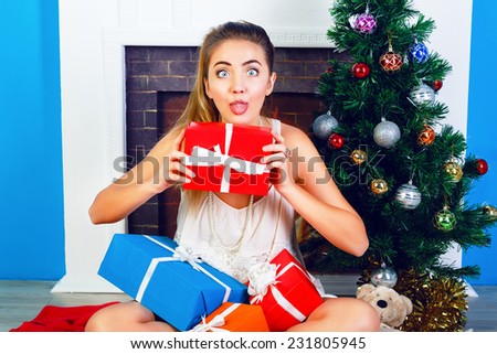Indoor bright portrait of funny young blonde girl sitting at home near New Year tree and opening Christmas presents. Show tongue and making funny faces.