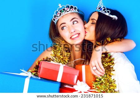 Funny lifestyle portrait of two pretty best friend girls wearing carnival crowns and tinsel , holding bright gifts and presents. Having fun at kiss on cheek in holiday night party.