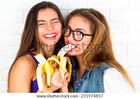 Two sisters eating bananas and having crazy funny time, smiling screaming and laughing. Wearing hipster sunglasses and trendy seductive make up.