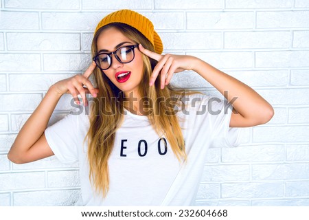 Fashion studio portrait of pretty young hipster blonde woman with bright sexy make up and glasses , wearing stylish urban t shirt and hat, White urban wall background.
