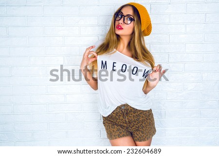 Fashion portrait of young pretty hipster woman with blonde hairs and big full bright lips wearing stylish outfit, denim swag shorts hat glasses and t shirt with funny print.urban wall background.