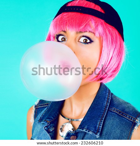 Close up funny fashion portrait of cheerful woman inflating the bubble gum, wearing swag hat, hot pink party wig, denim vest and stylish trendy diamond necklace. Bright sweet colors, mint background.