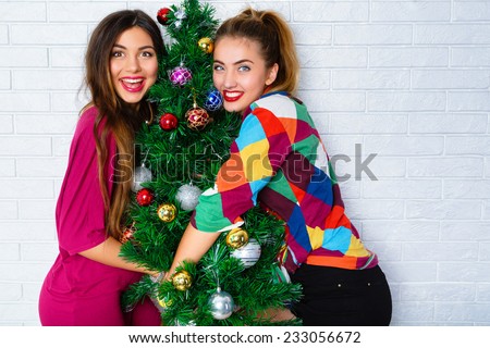 Close up lifestyle indoor portrait of two young woman posing near decorated Christmas tree, at New Year eve. Smiling having fun, ready for celebration. Bright holiday image of best friends.
