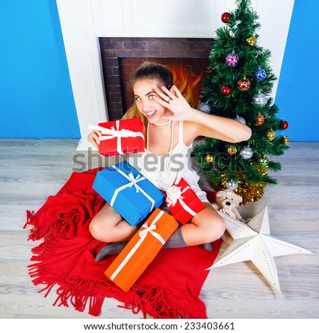 Lifestyle portrait of pretty happy blonde woman opening her presents in New year eve, wearing cute dress and sitting near fireplace and decorated Christmas tree. Cozy home atmosphere.