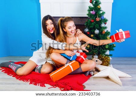 Two pretty best friends girls opening Christmas presents near fireplace and decorated New Year tree. Having fun together at winter holiday time.